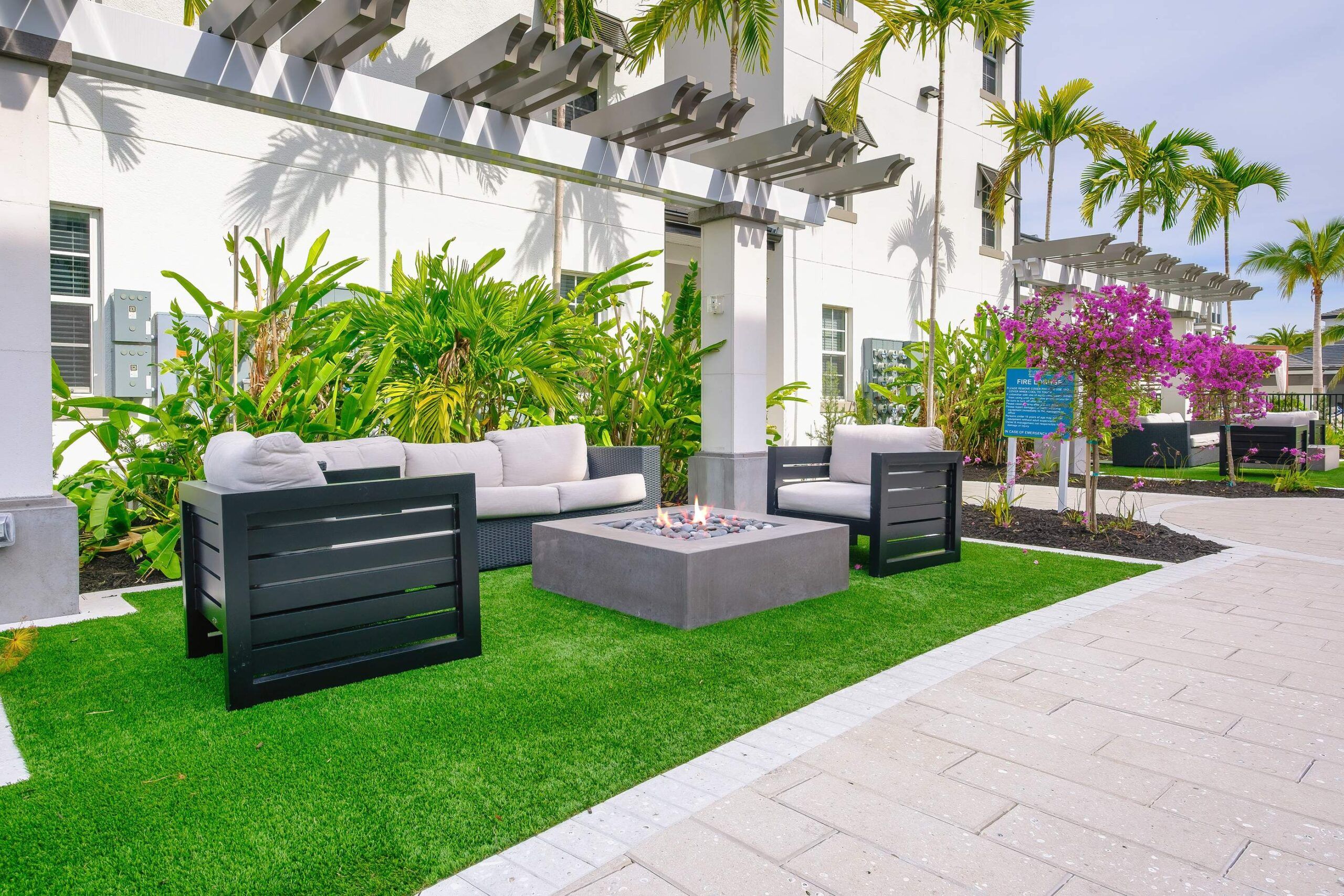 Inspira Naples Outdoor patio with modern seating, a fire pit, lush green plants, and palm trees next to a building.