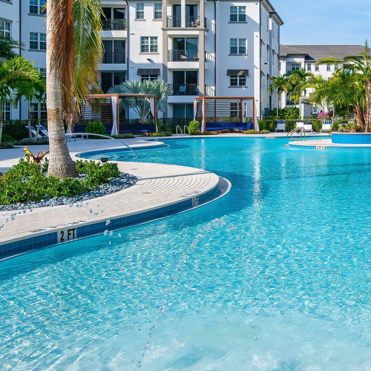 Inspira Naples Large apartment complex with balconies overlooks a clear blue swimming pool, palm trees, and sun loungers.