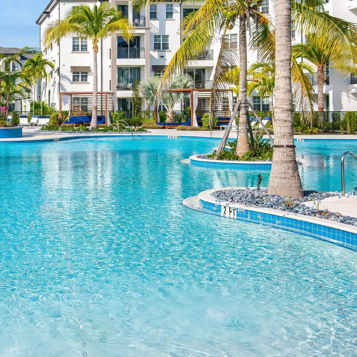 Inspira Naples Sunny outdoor pool with clear blue water, palm trees, and a modern white apartment building in the background.