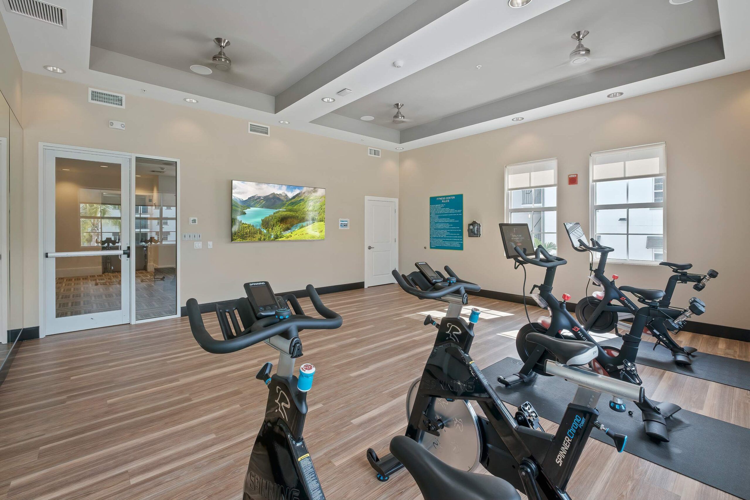 Inspira Naples Modern gym room with several stationary bikes, large windows, wooden floor, and a wall-mounted TV.